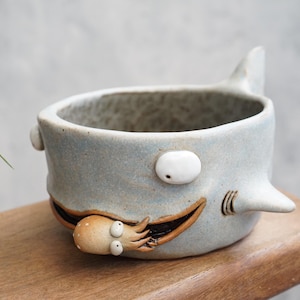 May include: A ceramic shark-shaped planter with a light blue glaze. The planter features a fin, eyes, and gills, and a small octopus figure emerging from the shark's mouth. The planter is sitting on a wooden surface.