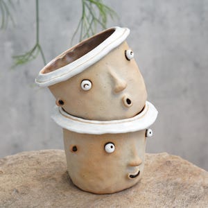 May include: Two stacked ceramic planters with whimsical face designs. The planters are beige with white accents around the rim and eyes. The faces have surprised expressions with open mouths and bulging eyes. These unique planters add a touch of art to any space.