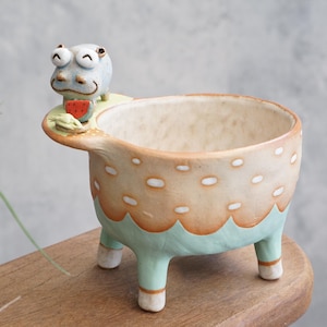 May include: A whimsical ceramic planter with a light blue hippo figure holding a slice of watermelon. The planter has a beige body with white polka dots, an orange trim, and mint green legs. A unique decorative item.