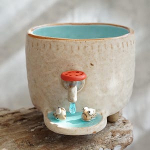 Puede incluir: Una taza de cerámica caprichosa con un exterior beige claro y un interior turquesa. Presenta un grifo rojo con un chorro de agua y dos pequeños peces en una piscina. La taza descansa sobre una superficie de madera.