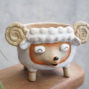 May include: A ceramic sheep-shaped planter with a cream-colored body, tan face, and curly horns. The planter has a smiling expression and is supported by three small legs. The interior is a light brown color.