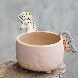 May include: A light pink ceramic planter featuring a sculpted unicorn head with a rainbow-striped mane and a matching rainbow-striped tail. The bowl has a textured surface, a tan interior, and a decorative gold-colored triangular pattern at the base.