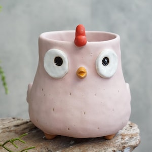 May include: A light pink ceramic planter shaped like a chicken. The planter features two large white circular eyes with black pupils, a small yellow beak, and a bright red comb on top. The body has a textured surface with small indentations.