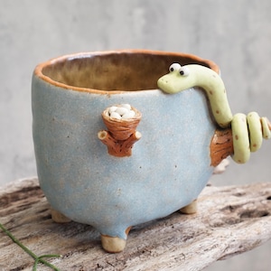 May include: A quirky ceramic plant pot in light blue, with a green snake handle and a small nest containing white objects. The pot has a brown rim and three small feet. A unique decorative item, ideal for a home.