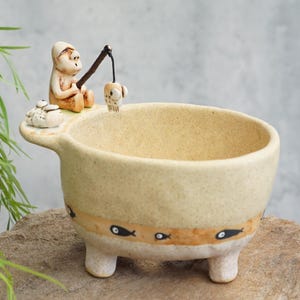 Puede incluir: Un cuenco de cerámica beige con tres patas cortas y textura moteada. Una pequeña figura esculpida se sienta en el borde sosteniendo una caña de pescar con un pez colgando. Una banda horizontal en el centro presenta iconos de peces negros pintados.