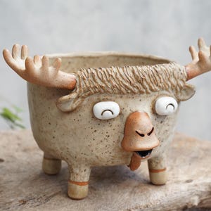 Puede incluir: Una maceta de cerámica beige moteada con forma de alce, con una melena texturizada, dos astas prominentes y un rostro con dos ojos blancos, una nariz marrón y una pequeña boca abierta. El recipiente se apoya sobre cuatro patas cortas.