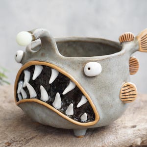 May include: A grey ceramic mug shaped like an anglerfish, featuring a wide mouth with sharp white teeth, two protruding eyes, a small white lure on a stalk, and three tan-coloured fins on the side. The interior is dark and smooth.