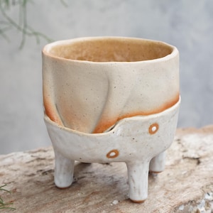 Puede incluir: Una maceta de cerámica hecha a mano con un acabado moteado blanquecino. La parte superior es de color beige claro con una banda rojo-anaranjada. La maceta tiene tres patas cortas y dos pequeños detalles decorativos redondos. La maceta está sobre un trozo de madera.
