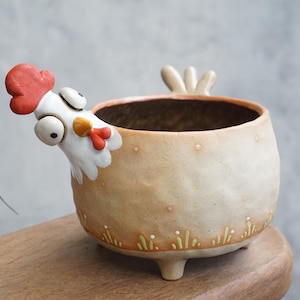 Puede incluir: Maceta de cerámica hecha a mano con forma de gallina. La maceta blanquecina presenta una cabeza de gallina con cresta roja y pico amarillo. La maceta tiene tres patas pequeñas y un diseño decorativo de hierba en la base.