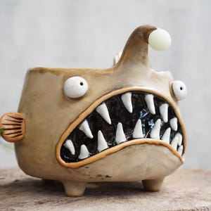 May include: A beige ceramic planter shaped like an anglerfish with a wide mouth, sharp white teeth, and a black interior. It features two protruding white eyes, a small side fin, and a lure extending from the top with a round white bulb.