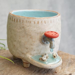 May include: A ceramic planter with a textured off-white exterior and a light blue interior. It features a decorative red and blue faucet handle on the side, with a small blue pool at the base containing three small sculpted fish figures.