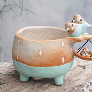 Puede incluir: Una maceta de cerámica caprichosa con un exterior marrón claro y naranja, acentuada con una base y patas verde menta. Una figura de pájaro se posa en el borde, sosteniendo una pequeña bandeja con pajaritos. Un artículo decorativo único.