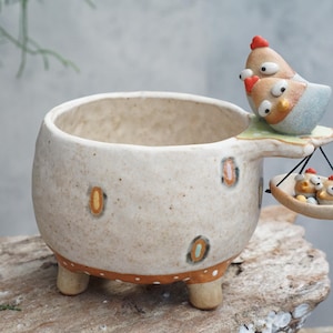 以下が含まれることがあります： 淡いベージュ仕上げの、気まぐれなセラミックプランター。茶色とオレンジ色のアクセントで飾られています。プランターには、2羽の成鶏と2羽のひよこが入った小さなトレイを含む、装飾的な鶏の家族が付いたハンドルが付いています。