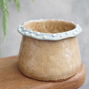 Puede incluir: Jarrón de cerámica hecho a mano, con cuerpo marrón claro y borde azul claro. El borde está decorado con pequeños puntos blancos. El jarrón está sobre una superficie de madera.