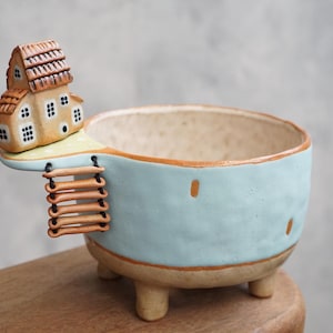 May include: A whimsical ceramic planter with a light blue exterior and a beige interior. A miniature house with a brown roof and windows is attached to the side, along with a ladder. The planter rests on three small legs.