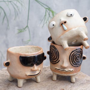 Puede incluir: Tres macetas de cerámica caprichosas con caras humanas. Una lleva gafas de sol, otra tiene ojos en espiral y la tercera tiene la nariz mocosa. Cada maceta es de color beige con detalles en negro y marrón y tiene patas cortas.