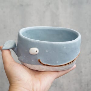 Puede incluir: Un cuenco de cerámica azul claro con forma de ballena, que presenta un pequeño ojo blanco, una línea marrón curva para la boca y una aleta caudal en el lateral. El cuenco tiene una textura moteada, una base blanca y un borde redondeado.