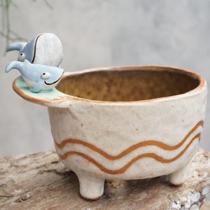 Puede incluir: Una maceta de cerámica con acabado beige claro y tres patas pequeñas. La maceta presenta dos figuras de ballenas azules en el borde y patrones de ondas naranjas alrededor del cuerpo. El interior es de color marrón dorado.