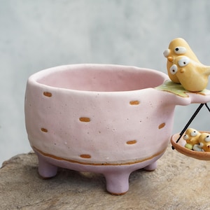 May include: A light pink ceramic planter with a textured surface featuring small brown dashes. A yellow bird figure sits on the rim, holding a small hanging scale with two tiny yellow bird figures inside, supported by thin black strings.