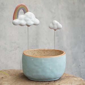 Puede incluir: Una maceta de cerámica azul claro con interior moteado y borde de terracota. Dos varillas metálicas delgadas se extienden hacia arriba desde la maceta, cada una rematada con una nube de cerámica blanca. Una nube tiene un arcoíris colorido.