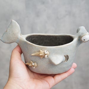 Puede incluir: Un cuenco de cerámica gris claro con forma de tiburón martillo, con una aleta caudal en un lado y una cabeza con ojos saltones en el otro. El exterior presenta dos pequeñas figuras de tiburón adheridas y hendiduras branquiales talladas.