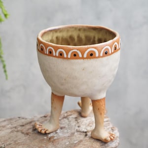 May include: A ceramic planter with a cream-colored, textured body and three legs shaped like human feet. The rim features a decorative orange band with white arches and black dots. The interior of the vessel has a mottled brown and tan glaze.