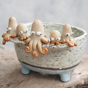May include: A ceramic planter shaped like a bathtub with five small, beige, tentacled octopus figures perched on the rim. The tub has a speckled off-white upper section, a brown band, and a light blue base with four short, sturdy legs.