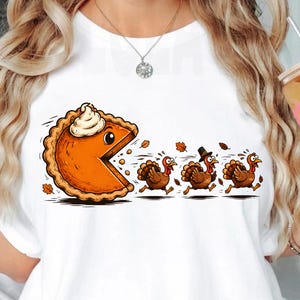 May include: A white t-shirt featuring a graphic of a large orange pumpkin pie slice with a dollop of whipped cream chasing three brown turkeys across the front. The turkeys are running away from the pie, with one wearing a black pilgrim hat.