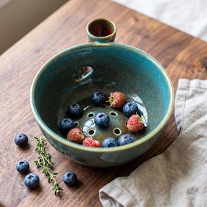 May include: A round, teal ceramic berry bowl with a handle and drainage holes at the base. The bowl contains fresh blueberries and raspberries. A sprig of green thyme and additional loose blueberries are placed nearby on the surface.
