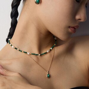 May include: A gold-toned jewelry set consisting of a layered necklace and a single stud earring. The necklace features a strand of small green beads and white pearls, paired with a delicate gold chain holding a small oval green stone pendant.
