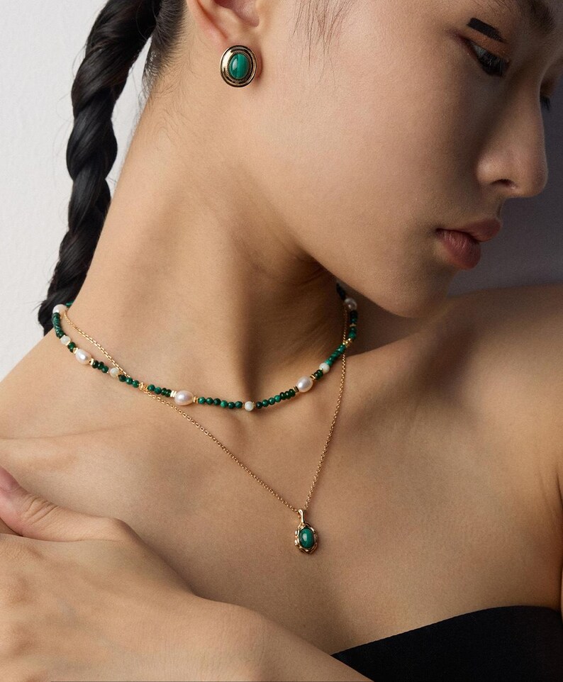 May include: A gold-toned jewelry set consisting of a layered necklace and a single stud earring. The necklace features a strand of small green beads and white pearls, paired with a delicate gold chain holding a small oval green stone pendant.