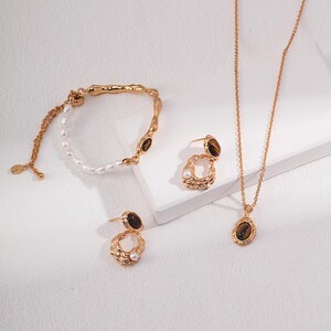 May include: A gold-toned jewelry set consisting of a bracelet, a pair of earrings, and a necklace. Each piece features a textured metal finish, small white pearls, and oval-shaped brown tiger eye gemstones set in gold-colored metal frames.