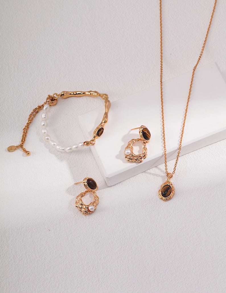 May include: A gold-toned jewelry set consisting of a bracelet, a pair of earrings, and a necklace. Each piece features a textured metal finish, small white pearls, and oval-shaped brown tiger eye gemstones set in gold-colored metal frames.