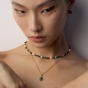 May include: A layered necklace set featuring a gold chain with a green stone pendant and a beaded choker with small green beads and white pearls. Matching gold earrings with oval green stones and a gold ring with green and orange stones.