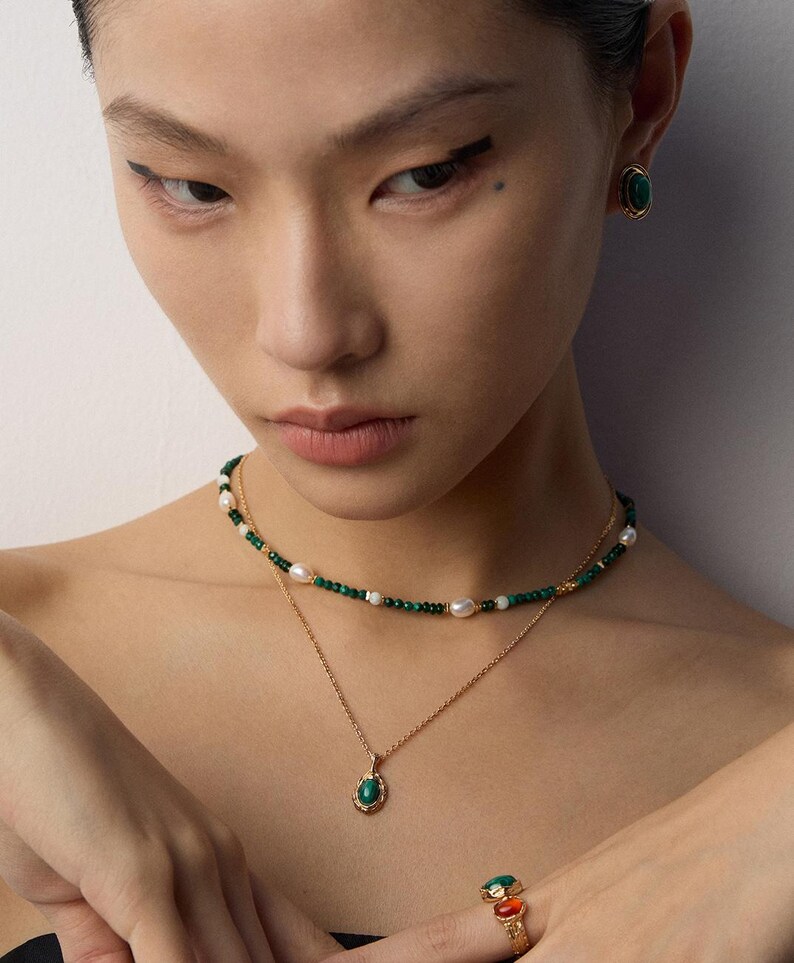 May include: A layered necklace set featuring a gold chain with a green stone pendant and a beaded choker with small green beads and white pearls. Matching gold earrings with oval green stones and a gold ring with green and orange stones.