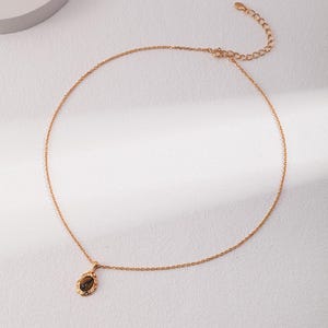 May include: A gold-toned chain necklace featuring a single oval pendant with a brown, striped tiger's eye gemstone set in a textured metal frame. The necklace includes a delicate link chain and an adjustable extender with a small circular tag.