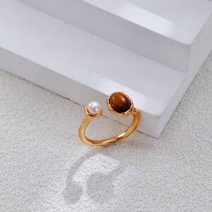 May include: An open-style gold-toned ring featuring a small white pearl on one end and a larger, oval-shaped brown tiger's eye gemstone on the other. The band has a smooth, polished finish and a slightly irregular, organic shape.