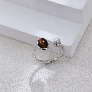 May include: A silver-toned open ring featuring a polished oval brown tiger's eye gemstone on one end and a small, round white pearl on the other. The band has a smooth, slightly irregular metallic finish, creating a distinct contrast between the stones.