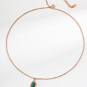 May include: A rose gold-toned necklace featuring a delicate chain and a single pendant. The pendant consists of an oval-shaped, dark green stone set within a textured, organic-looking rose gold metal frame. The chain includes an adjustable extender.