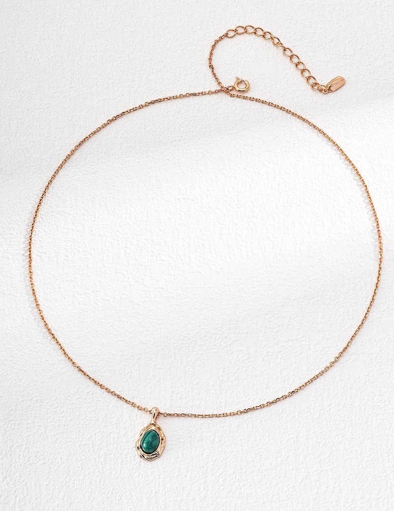 May include: A rose gold-toned necklace featuring a delicate chain and a single pendant. The pendant consists of an oval-shaped, dark green stone set within a textured, organic-looking rose gold metal frame. The chain includes an adjustable extender.