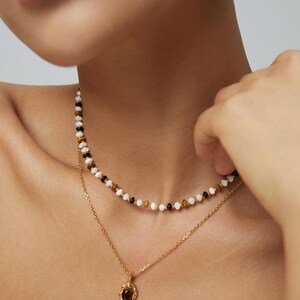 May include: A layered necklace set consisting of a short beaded strand featuring alternating white, black, and brown beads, and a longer gold-toned chain with a small oval pendant containing a dark brown stone set in a textured gold frame.