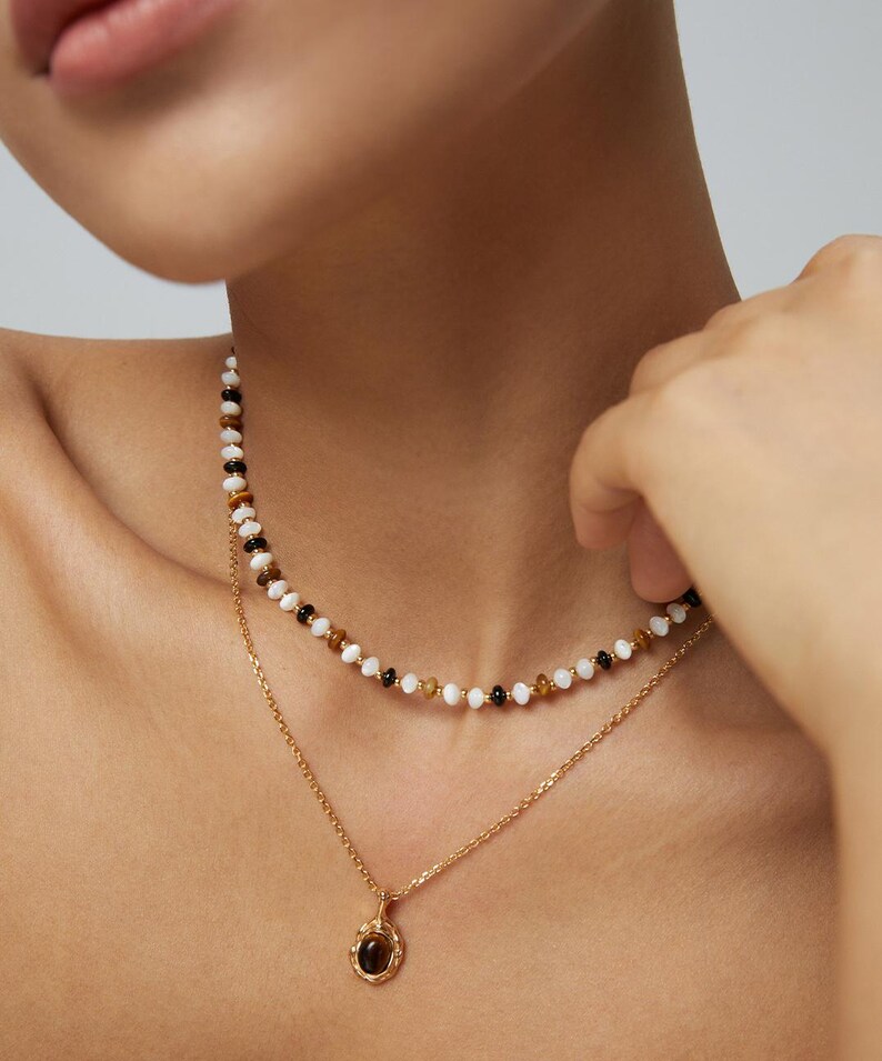 May include: A layered necklace set consisting of a short beaded strand featuring alternating white, black, and brown beads, and a longer gold-toned chain with a small oval pendant containing a dark brown stone set in a textured gold frame.