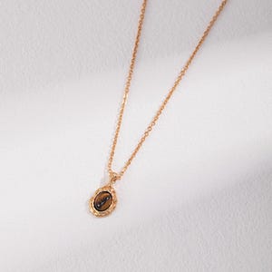 May include: A gold-toned necklace featuring a delicate chain and an oval pendant. The pendant contains a smooth, brown, chatoyant tiger's eye gemstone set within a textured, scalloped gold-colored metal frame that adds detail to the piece.