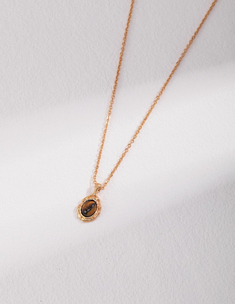 May include: A gold-toned necklace featuring a delicate chain and an oval pendant. The pendant contains a smooth, brown, chatoyant tiger's eye gemstone set within a textured, scalloped gold-colored metal frame that adds detail to the piece.