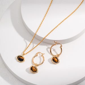 May include: A gold-toned jewelry set consisting of a necklace and a pair of hoop earrings. Each piece features a smooth, oval-shaped tiger's eye gemstone set in a textured, irregular gold-colored metal frame. The necklace has a twisted chain.
