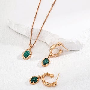 May include: A gold-toned jewelry set consisting of a necklace and two hoop earrings. Each piece features a polished, oval-shaped green malachite stone set within a textured, organic-looking gold metal frame with a warm, metallic finish.