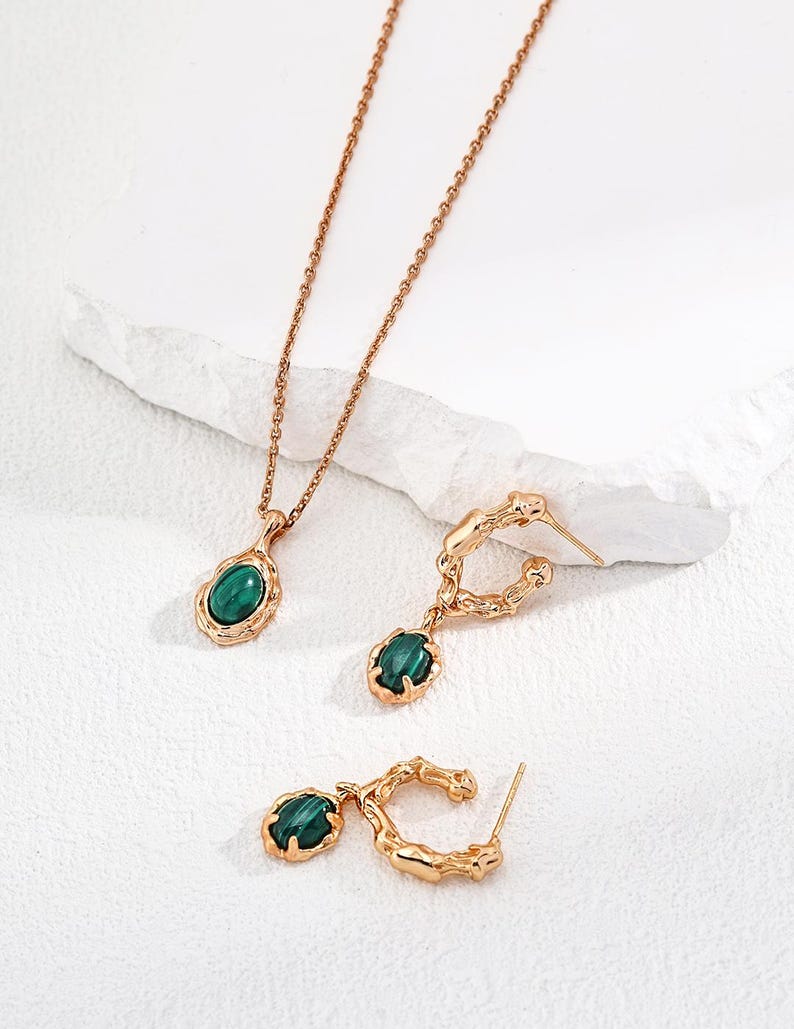 May include: A gold-toned jewelry set consisting of a necklace and two hoop earrings. Each piece features a polished, oval-shaped green malachite stone set within a textured, organic-looking gold metal frame with a warm, metallic finish.