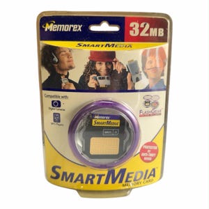 May include: A Memorex 32MB SmartMedia memory card in its original clear plastic blister packaging. The card is black with a gold contact area and is housed in a purple circular protective case. The packaging features product branding and text.