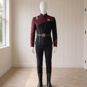 Könnte beinhalten: Eine Star-Trek-Uniform bestehend aus einer schwarzen und kastanienbraunen Jacke mit Stehkragen, einem goldenen Starfleet-Abzeichen auf der Brust und einem schwarzen Gürtel mit runder goldener Schnalle. Das Outfit umfasst eine schwarze Hose mit kastanienbraunen Seitenstreifen.