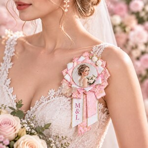 Könnte beinhalten: Eine rosafarbene und weiße Stoffrosetten-Brosche mit einem runden Fotoeinsatz einer Person im Brautkleid, einem kleinen Schmetterlingsakzent und zwei herabhängenden Bändern. Das linke Band zeigt die rosa gestickten Buchstaben M & L vertikal.